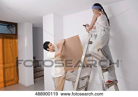 Stock Photo - Young woman hammering nail on the wall with a young man holding a picture frame. Fotosearch