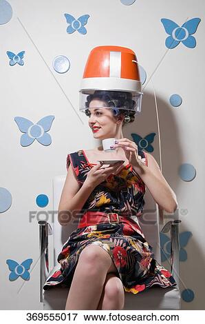 Stock Photo - Young woman sitting under a hair dryer and drinking tea. Fotosearch