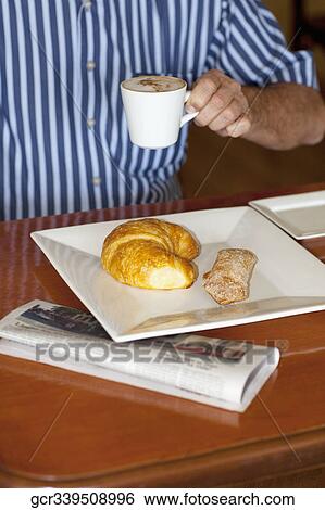 Stock Photograph - Mid section view of a man drinking coffee with a croissant and cracker in front of him. Fotosearch