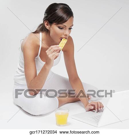 Picture - Woman eating a snack and working on a laptop. Fotosearch