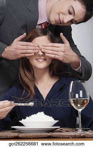Stock Image - Woman eating rice and a man covering her eyes. Fotosearch