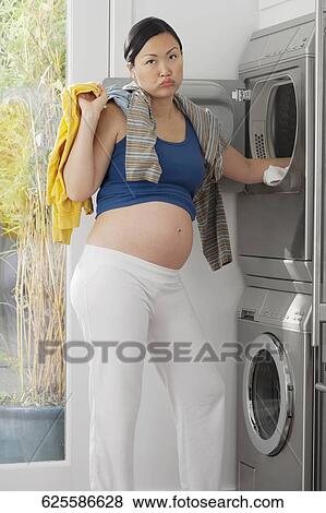 Stock Photo - Annoyed pregnant woman holding clothes in front of a washing machine. Fotosearch