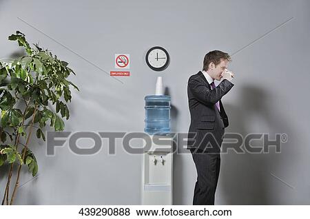 Businessman drinking water near a water cooler View Large Photo Image Stock Photo - Businessman drinking water near a water cooler. Fotosearch