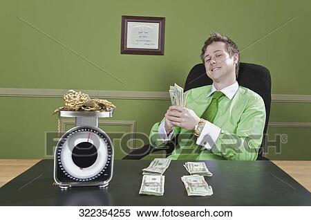 Stock Photography - Businessman holding currency notes with gold jewelry on a weight scale in front of him. Fotosearch