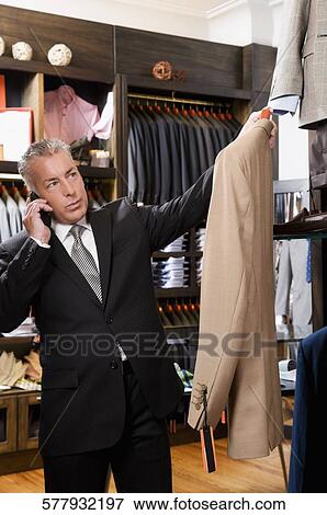 Businessman selecting a suit and talking on a mobile phone in a clothing store View Large Photo Image Stock Photo - Businessman selecting a suit and talking on a mobile phone in a clothing store. Fotosearch
