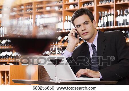 Stock Image - Businessman talking on a mobile phone and using a laptop in a bar. Fotosearch