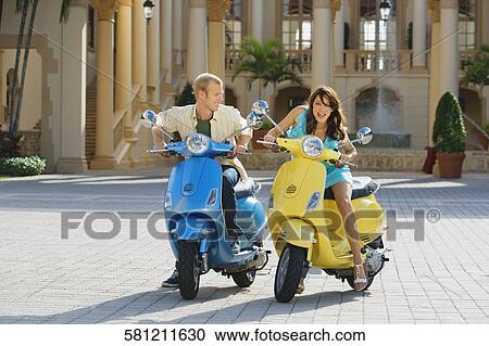 Stock Image - Couple sitting on mopeds, Biltmore Hotel, Coral Gables, Florida, USA. Fotosearch