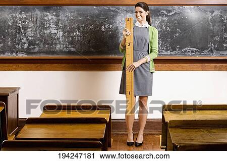 Stock Image - Female teacher holding a ruler in a classroom and smiling. Fotosearch