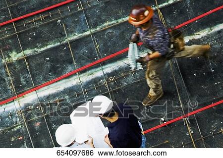 Stock Photograph - High angle view of two architects standing with a construction worker at a construction site. Fotosearch