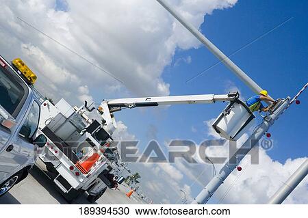 Stock Image - Low angle view of a maintenance engineer on a mobile crane repairing a security camera on a pole. Fotosearch