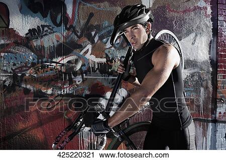 Stock Image - Man carrying a bicycle on his shoulders in front of a graffiti covered wall. Fotosearch