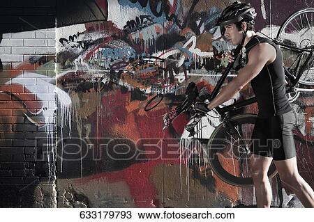 Stock Image - Man carrying a bicycle on his shoulders in front of a graffiti covered wall. Fotosearch