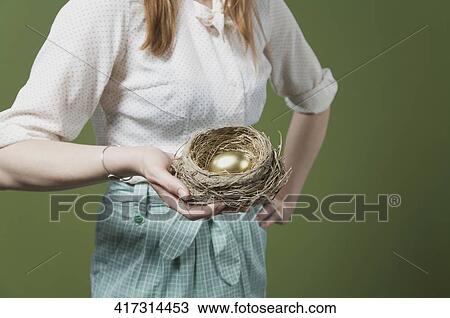 Stock Image - Mid section view of a woman holding a golden egg in the nest. Fotosearch
