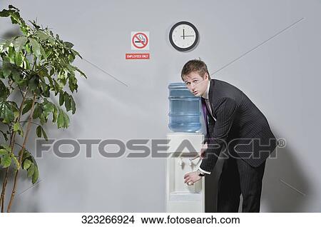 Picture - Portrait of a businessman pouring water into a glass from a water cooler. Fotosearch
