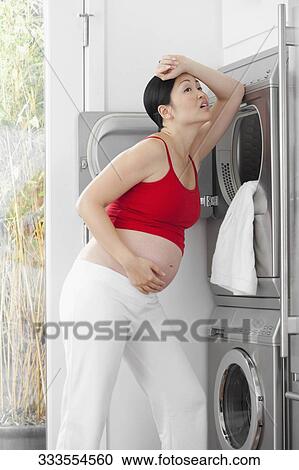 Stock Image - Pregnant woman looking tired in front of a washing machine. Fotosearch