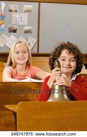 Schoolboy holding a bell with a schoolgirl sitting behind him in a classroom View Large Photo Image Stock Photography - Schoolboy holding a bell with a schoolgirl sitting behind him in a classroom. Fotosearch