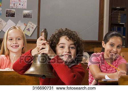 Stock Image - Schoolboy holding a bell with two schoolgirls sitting behind him in a classroom. Fotosearch