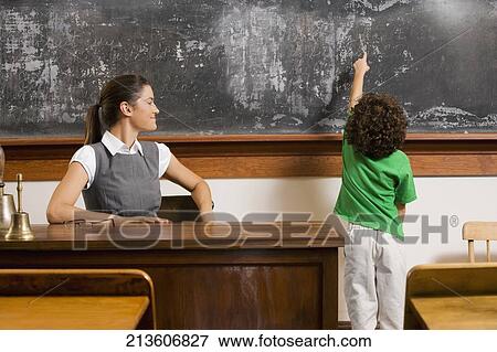 Stock Photo - Schoolboy pointing at a blackboard with a teacher in a classroom. Fotosearch