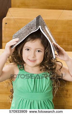 Schoolgirl holding a book on her head in a classroom View Large Photo Image Stock Photo - Schoolgirl holding a book on her head in a classroom. Fotosearch