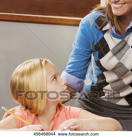 Schoolgirl studying in a classroom with her teacher smiling behind her View Large Photo Image Picture - Schoolgirl studying in a classroom with her teacher smiling behind her. Fotosearch