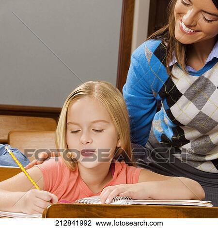 Schoolgirl studying in a classroom with her teacher smiling behind her View Large Photo Image Stock Image - Schoolgirl studying in a classroom with her teacher smiling behind her. Fotosearch