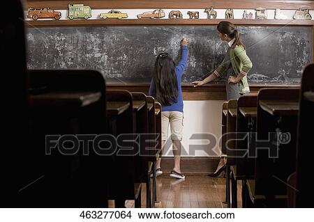 Schoolgirl writing on a blackboard in a classroom with her teacher standing beside her View Large Photo Image Picture - Schoolgirl writing on a blackboard in a classroom with her teacher standing beside her. Fotosearch