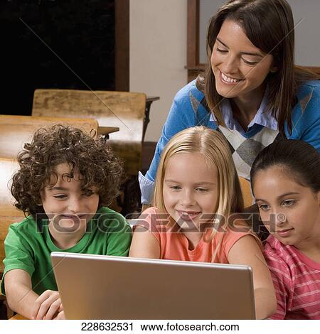 Stock Image - Students working on a laptop in a classroom with their teacher behind them. Fotosearch