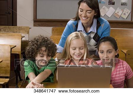 Stock Photo - Students working on a laptop in a classroom with their teacher behind them. Fotosearch