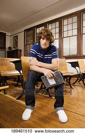 Stock Image - Teenage boy holding a book and sitting in a classroom. Fotosearch