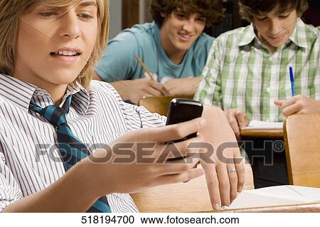 Stock Image - Teenage boy text messaging on a mobile phone in a classroom. Fotosearch