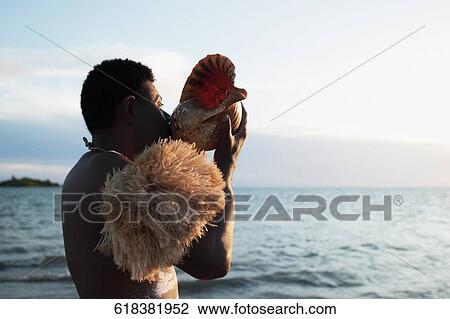 Stock Image - Tribesman blowing a cockleshell on the beach. Fotosearch