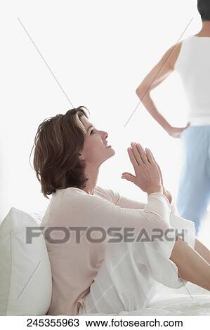 Stock Image - Woman sitting on the bed with her hands folded with a man standing in the background. Fotosearch