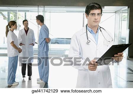 Stock Photo - Doctor holding a clipboard with his colleagues discussing in the background. Fotosearch