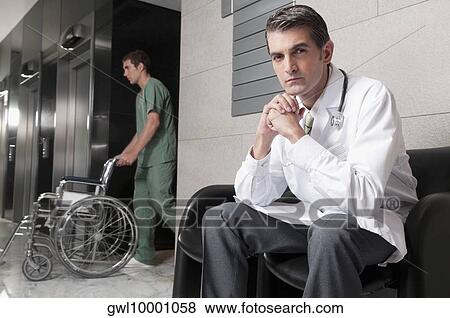 Doctor sitting on a chair with male nurse carrying a wheelchair View Large Photo Image Stock Photo - Doctor sitting on a chair with male nurse carrying a wheelchair. Fotosearch