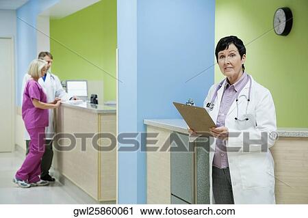 Stock Image - Female doctor holding a clipboard with a doctor and nurse in the background. Fotosearch