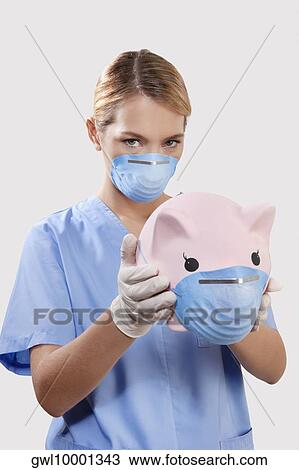 Stock Image - Female doctor wearing a flue mask and holding a piggy bank. Fotosearch