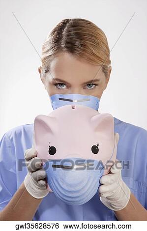 Stock Photograph - Female doctor wearing a flue mask and showing a piggy bank. Fotosearch