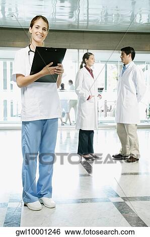 Female nurse holding a clipboard with her colleagues discussing in the background View Large Photo Image Stock Photograph - Female nurse holding a clipboard with her colleagues discussing in the background. Fotosearch