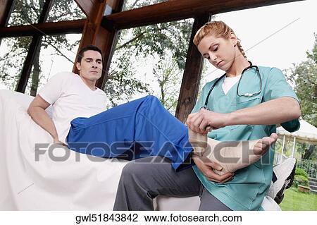 Stock Image - Female nurse rolling crepe bandage on a man's foot. Fotosearch