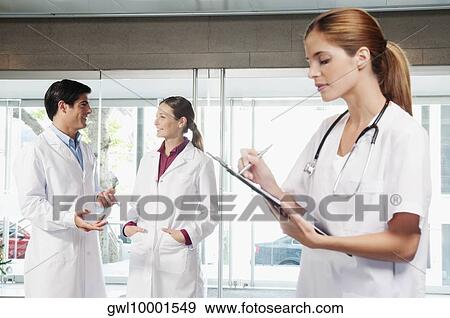 Female nurse writing on a clipboard with her colleagues discussing in the background View Large Photo Image Stock Photo - Female nurse writing on a clipboard with her colleagues discussing in the background. Fotosearch