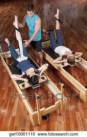 Stock Image - Male instructor assisting women in a gym. Fotosearch