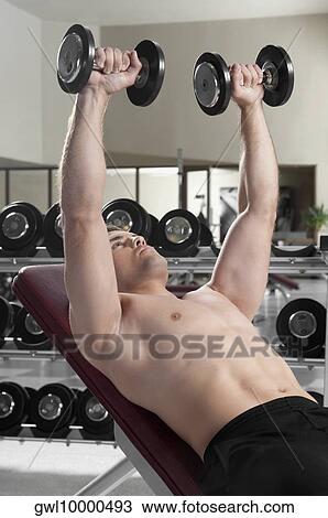 Stock Image - Man lying on a weight bench and exercising with dumbbells. Fotosearch