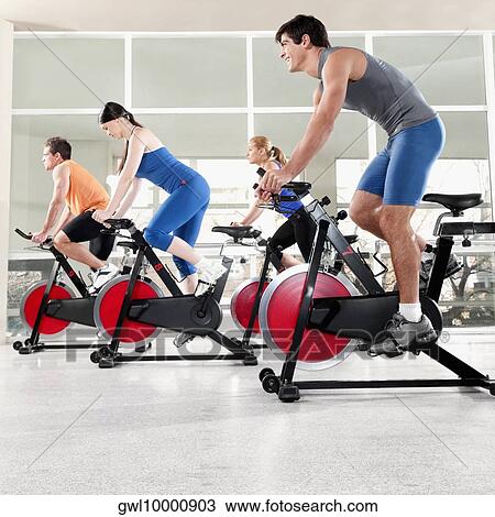 Stock Image - Men and women working out on exercise bikes in a gym. Fotosearch