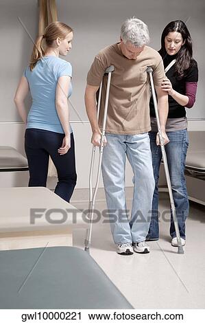 Stock Image - Physiotherapist assisting a patient in walking with crutches. Fotosearch