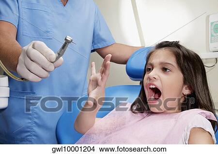 Picture - Scared girl looking at dental drill in a dentist's hand. Fotosearch