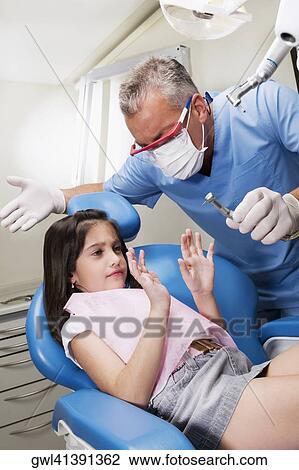 Stock Image - Scared girl looking at dental drill in a dentist's hand. Fotosearch