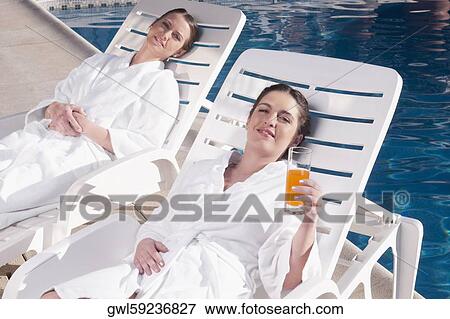 Stock Photo - Two women resting on lounge chairs at poolside. Fotosearch