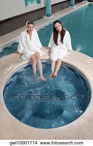 Picture - Two women sitting at the poolside with their feet soaking in water. Fotosearch