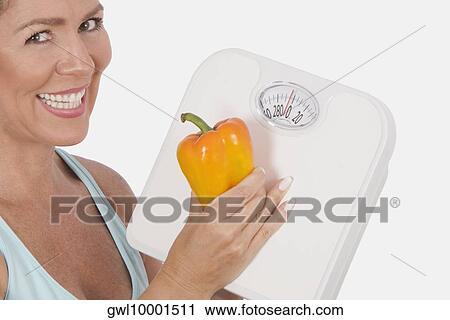 Stock Image - Woman holding a bathroom scale and an orange bell pepper and smiling. Fotosearch