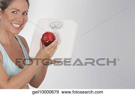 Woman holding a bathroom scale and an apple and smiling View Large Photo Image Stock Photo - Woman holding a bathroom scale and an apple and smiling. Fotosearch